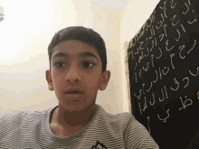a boy stands in front of a blackboard with arabic writing