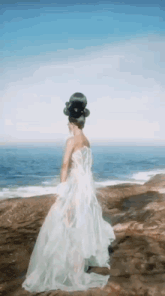a woman in a white dress and a hat is standing on a rocky cliff overlooking the ocean .