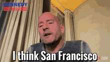 a bald man says i think san francisco in front of a yellow curtain