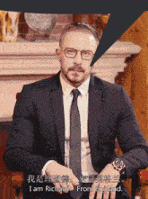 a man in a suit and tie is sitting in front of a fireplace and says " i am richard from england "