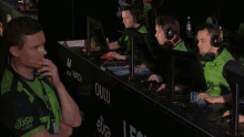 a man wearing a green elise shirt sits at a desk with other people