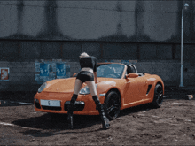 a woman wearing shorts with the number 60 on them stands in front of an orange car