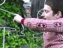 a man in a pink sweater is standing in front of a bush holding a stick .