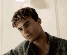 a young man with curly hair and a plaid shirt looks at the camera
