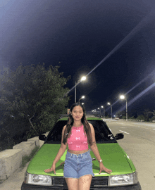 a woman in a pink top and denim shorts stands next to a green car