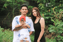 a man and a woman holding a baby in front of a lush green background