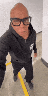 a bald man wearing glasses and a black hoodie stands in a hallway .