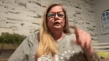 a woman wearing glasses and a gray shirt is making a gesture with her hand .