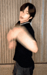a young man in a black shirt is dancing in front of a window with vertical blinds .