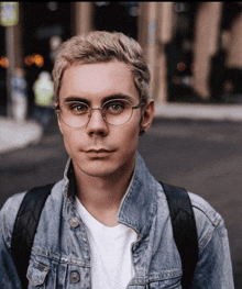 a man wearing glasses and a denim jacket has a backpack on his back