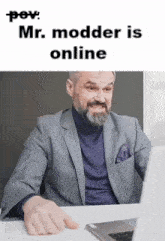 a man with a beard is sitting at a desk in front of a laptop computer with the caption " mr. modder is online "