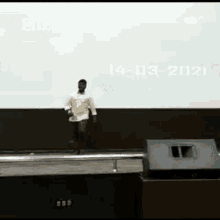 a man is standing on a stage in front of a large screen with the date of 14-03-2021 on it .
