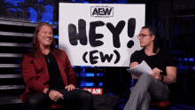 two men sitting in front of a sign that says hey