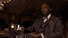 a man in a suit and bow tie is sitting at a table with a glass of whiskey in his hand .