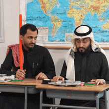 two men sit at desks in front of a world map with arabic writing