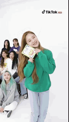 a girl in a green sweater is holding a book in front of a group of girls on tiktok live