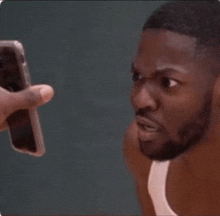 a man in a white tank top is looking at another man holding a cell phone .