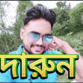 a man wearing sunglasses and a blue shirt is smiling in front of a green and yellow sign that says darkan