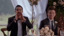 two men in tuxedos are sitting at a table with flowers and candles