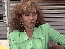 a woman in a green shirt is sitting in front of a window with blinds .