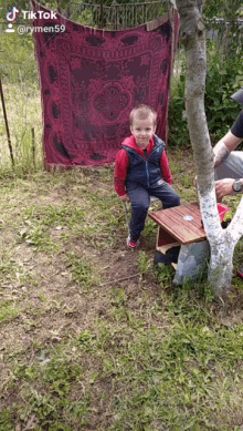 a little boy is sitting on a small table under a tree with a tik tok video behind him