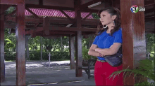 a woman in a blue shirt and red pants stands in front of a wooden structure with the number 28 on the bottom