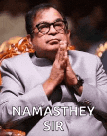 a man wearing glasses and a suit is sitting in a chair with his hands folded in prayer .