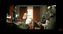 a group of men are standing around a man laying on a bed