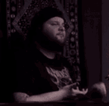 a man with a beard is wearing a black hat and a black shirt and is sitting in a dark room .