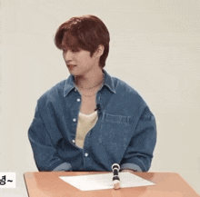 a young man in a denim shirt is sitting at a desk with a pen in his hand .