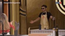 a man in an apron with the letter m on it is standing in front of a woman in a kitchen .