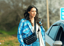 a woman wearing a blue plaid jacket and a black fanny pack walks towards a car