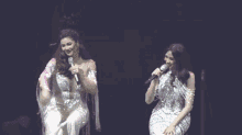 two women singing into microphones on a stage in front of a crowd