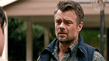 a man with a beard and a vest is standing in front of a house talking to another man .