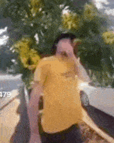 a man in a yellow shirt is standing in front of a white car and covering his nose .