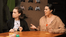 two women sit at a table in front of a wall with game controllers on it