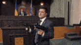 a man in a suit and tie is holding two guns in front of a podium with a ukrainian flag on it