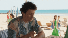 a man in a hawaiian shirt sits at a table by the beach