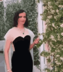 a woman in a black dress is standing in front of a bush with flowers .