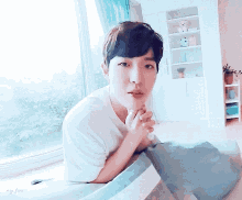 a young man in a white shirt is sitting on a bed in front of a window looking at the camera .
