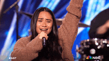 a woman is singing into a microphone with her arms in the air and a nbc logo in the background