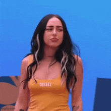 a woman in a yellow tank top with the word diesel on it is standing in front of a blue wall .
