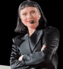 a woman in a black suit with a microphone on her head is sitting at a table with her arms crossed .