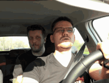 a man wearing glasses is driving a car with another man in the back seat