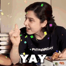 a woman is sitting at a table with a box of food and confetti falling around her and giving a thumbs up .