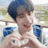 a young man in a plaid jacket makes a heart shape with his hands
