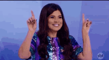a woman in a purple and blue sequined dress is pointing up with both hands .