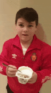 a boy wearing a red umbro jacket is eating yogurt with a spoon