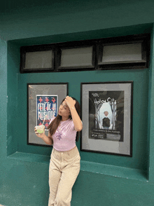 a woman leans against a green wall holding a drink in front of two framed posters one of which is titled hollow city