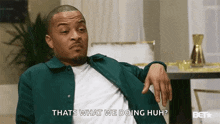 a man in a green jacket is sitting in a living room with his hand on his knee and talking to someone .